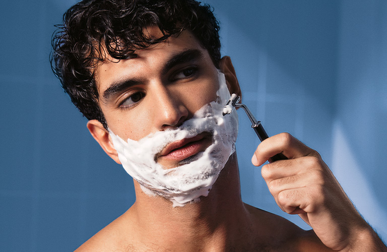 Foto de un hombre afeitándose con espuma y maquinilla. Fondo azul con sombra de luz. Se ve el torso descubierto, el rostro con espuma y la mano sosteniendo la afeitadora.